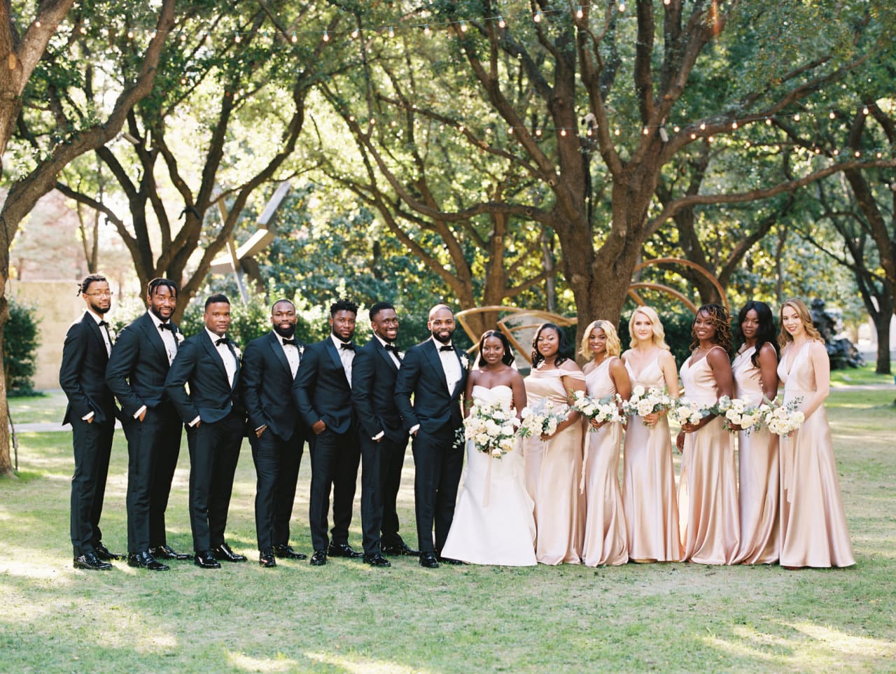 Full wedding party in black tie and champagne gowns beneath the trees